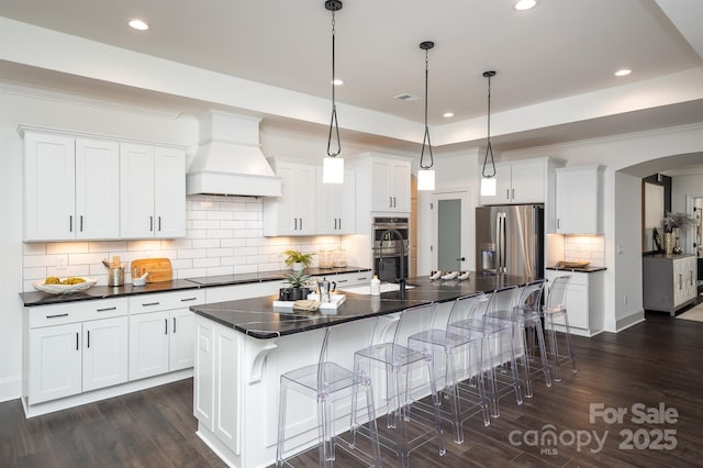 kitchen with appliances with stainless steel finishes, dark wood-type flooring, white cabinets, premium range hood, and a kitchen bar