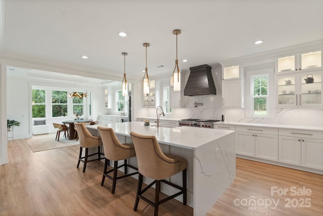 kitchen with an island with sink, light wood-style floors, premium range hood, white cabinetry, and a sink