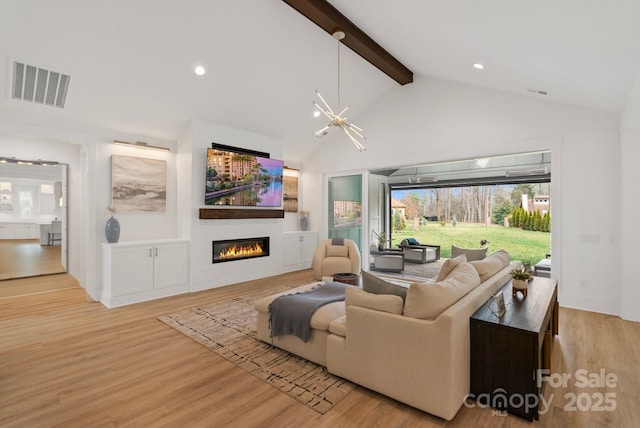 living area featuring visible vents, light wood-style floors, a glass covered fireplace, a chandelier, and beamed ceiling