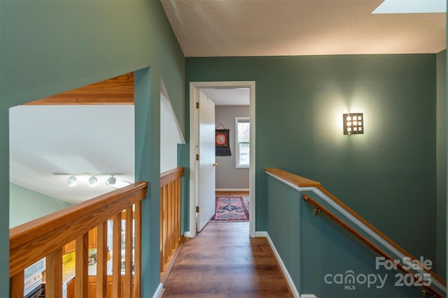 corridor featuring wood finished floors, a textured ceiling, track lighting, an upstairs landing, and baseboards