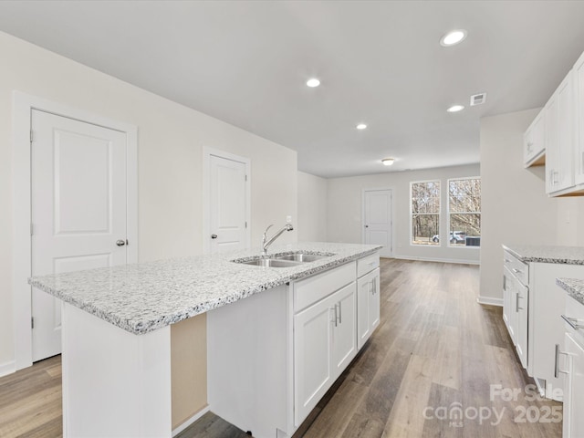 kitchen featuring an island with sink, light stone countertops, light wood-style floors, white cabinetry, and a sink