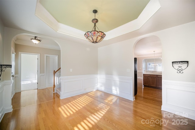 spare room with arched walkways, a wainscoted wall, stairway, a tray ceiling, and light wood-type flooring