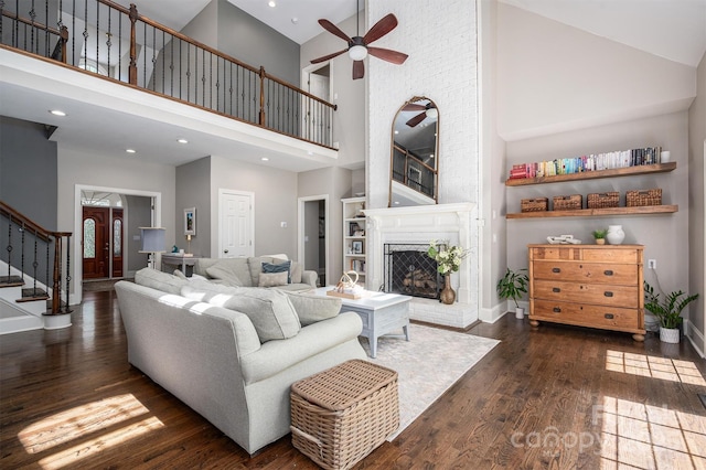 living room with dark wood-style flooring, a towering ceiling, a ceiling fan, a brick fireplace, and stairs