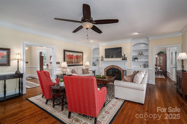 living room with built in shelves, dark wood-type flooring, ornamental molding, a brick fireplace, and baseboards