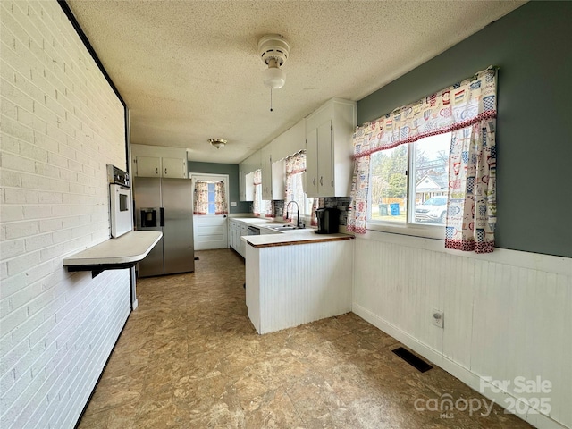 kitchen with a textured ceiling, a wainscoted wall, brick wall, visible vents, and stainless steel refrigerator with ice dispenser