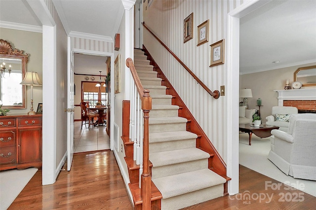 stairway with wallpapered walls, ornamental molding, wood finished floors, an inviting chandelier, and a brick fireplace