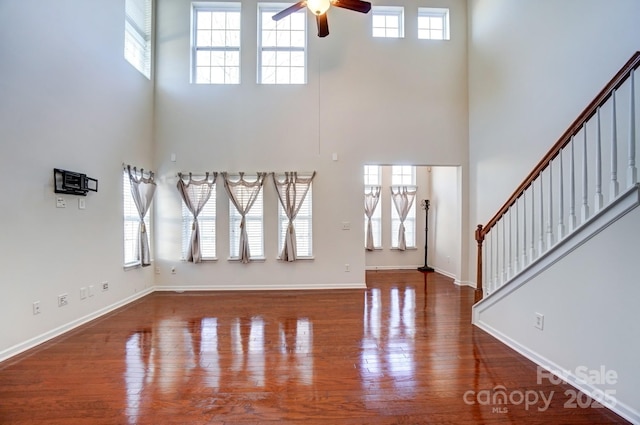 unfurnished living room featuring baseboards, ceiling fan, wood finished floors, a high ceiling, and stairs