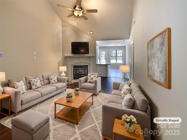 living area with high vaulted ceiling, a glass covered fireplace, a ceiling fan, and wood finished floors