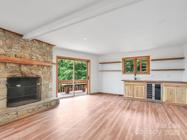 unfurnished living room with a stone fireplace, beverage cooler, light wood-type flooring, beamed ceiling, and indoor wet bar