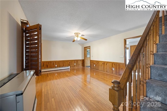 unfurnished living room featuring a textured ceiling, baseboard heating, wainscoting, and light wood-style flooring