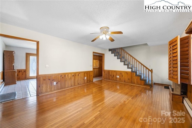 unfurnished living room featuring a textured ceiling, a ceiling fan, stairway, wainscoting, and hardwood / wood-style floors