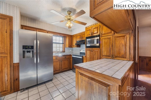 kitchen with brown cabinets, tile countertops, stainless steel appliances, under cabinet range hood, and wallpapered walls