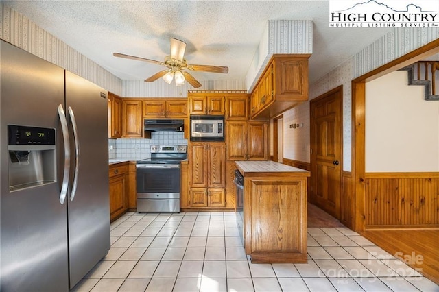 kitchen featuring wallpapered walls, a wainscoted wall, appliances with stainless steel finishes, a textured ceiling, and under cabinet range hood