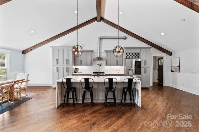 kitchen with a kitchen island with sink, gray cabinetry, light countertops, wall chimney range hood, and stainless steel refrigerator with ice dispenser