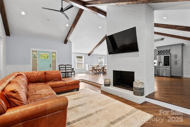 living room featuring a ceiling fan, dark wood-style floors, a wainscoted wall, beamed ceiling, and a brick fireplace