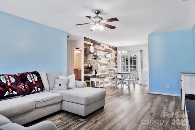 living room with an accent wall, wooden walls, wood finished floors, baseboards, and ceiling fan with notable chandelier