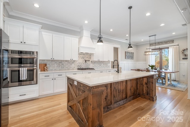 kitchen featuring stainless steel appliances, light countertops, ornamental molding, wainscoting, and premium range hood