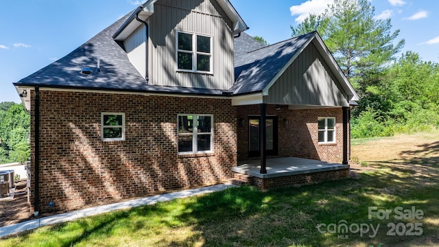 rear view of house with a patio, brick siding, a lawn, roof with shingles, and board and batten siding