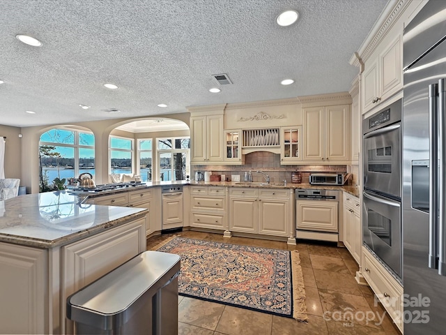 kitchen with stainless steel appliances, a sink, cream cabinetry, decorative backsplash, and glass insert cabinets