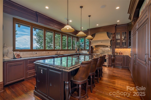 kitchen with custom exhaust hood, a center island with sink, decorative backsplash, dark wood finished floors, and glass insert cabinets