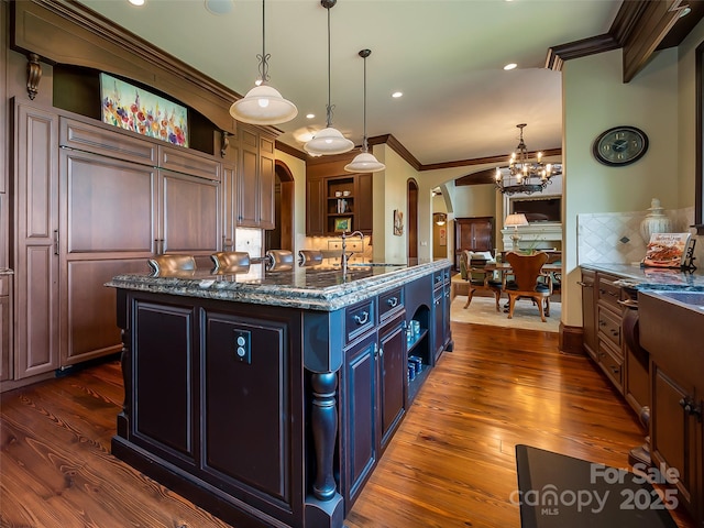 kitchen with arched walkways, ornamental molding, open shelves, an island with sink, and dark wood finished floors