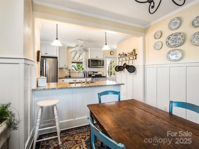 kitchen with white cabinets, a wainscoted wall, appliances with stainless steel finishes, light stone counters, and a sink