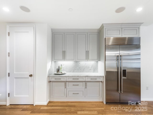 kitchen with stainless steel built in fridge, light stone countertops, gray cabinetry, light wood-type flooring, and backsplash