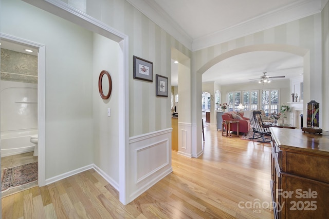 hallway with light wood-style floors, arched walkways, a wainscoted wall, and ornamental molding