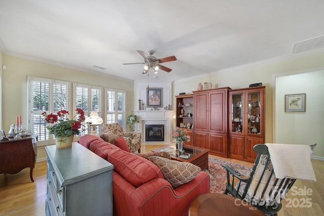 living area featuring crown molding, visible vents, a ceiling fan, a large fireplace, and light wood-type flooring