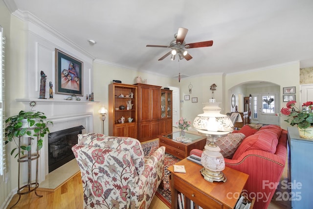 living room featuring arched walkways, a fireplace with raised hearth, a ceiling fan, ornamental molding, and light wood-type flooring