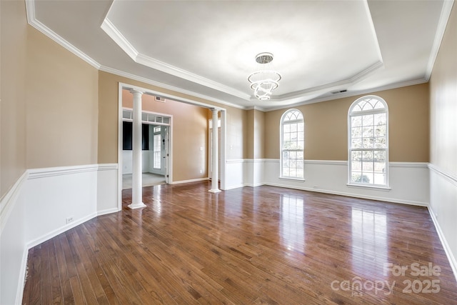 empty room with decorative columns, a tray ceiling, wood finished floors, and wainscoting