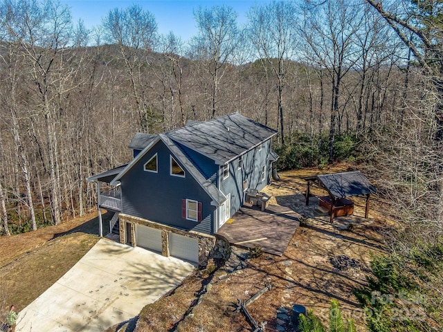 exterior space featuring a garage, roof with shingles, a wooded view, and concrete driveway