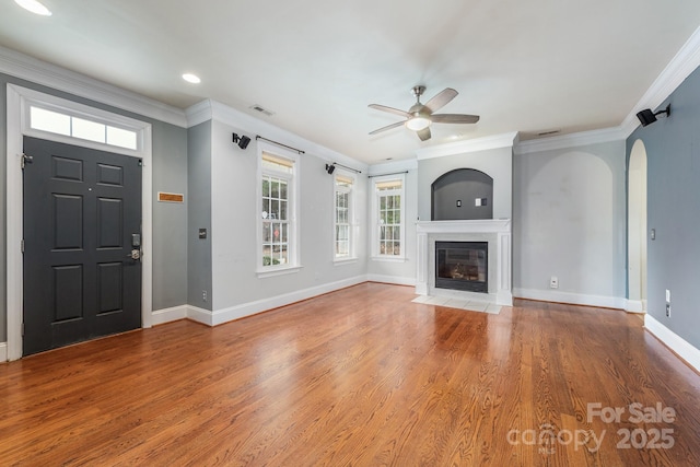 unfurnished living room with arched walkways, wood finished floors, a fireplace with flush hearth, baseboards, and crown molding