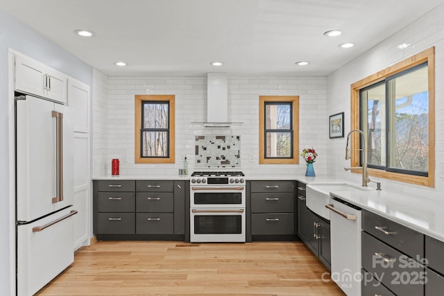 kitchen with decorative backsplash, premium appliances, wall chimney exhaust hood, light wood-style floors, and a sink