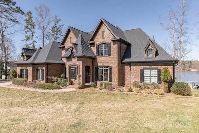 french country inspired facade with brick siding, roof with shingles, a front yard, and a water view