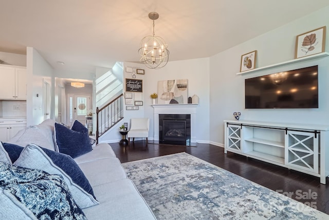 living area with baseboards, a glass covered fireplace, stairway, dark wood-style flooring, and a notable chandelier