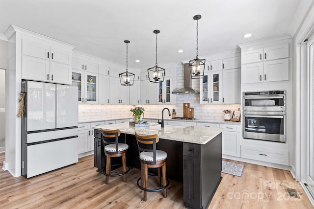 kitchen featuring tasteful backsplash, white cabinets, light wood-style flooring, stainless steel appliances, and wall chimney range hood