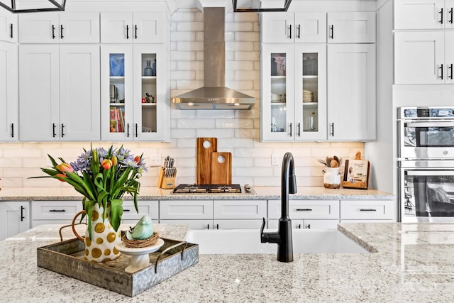 kitchen with white cabinetry, wall chimney range hood, appliances with stainless steel finishes, decorative backsplash, and glass insert cabinets