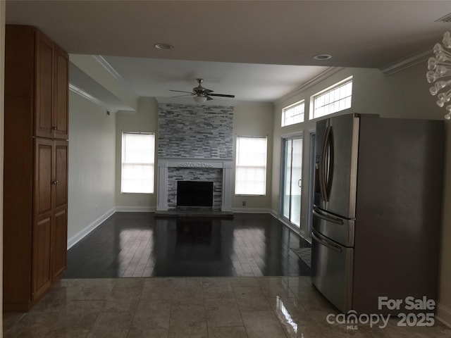 unfurnished living room with ceiling fan, a stone fireplace, ornamental molding, and baseboards