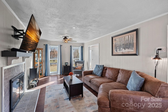 living area with ceiling fan, a textured ceiling, wood finished floors, a brick fireplace, and crown molding