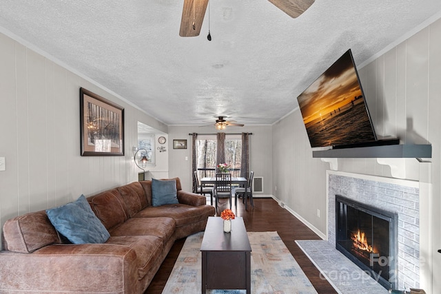 living area featuring dark wood-style flooring, crown molding, a ceiling fan, a brick fireplace, and a textured ceiling