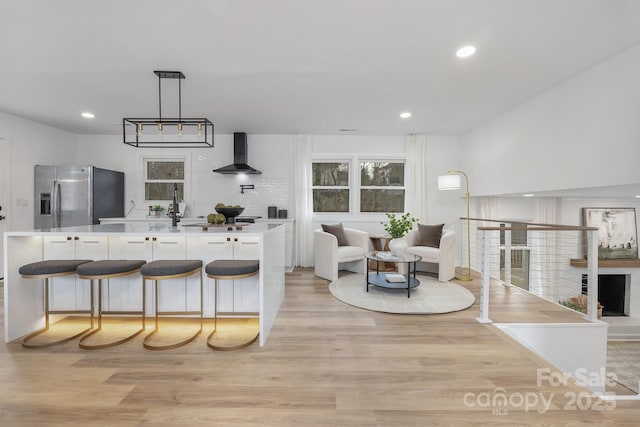 kitchen featuring hanging light fixtures, light countertops, light wood-type flooring, stainless steel refrigerator with ice dispenser, and wall chimney exhaust hood