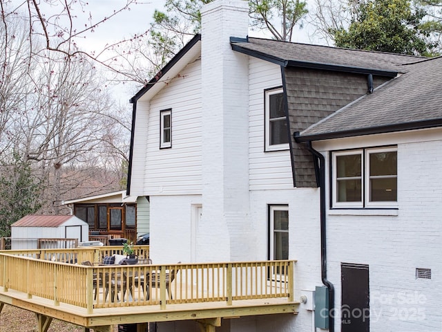 back of house with brick siding, roof with shingles, a chimney, a deck, and a shed