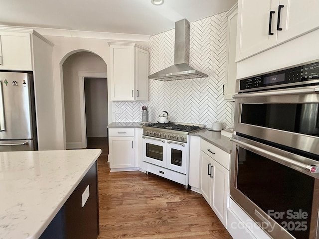 kitchen with light stone countertops, stainless steel appliances, wall chimney range hood, white cabinetry, and backsplash