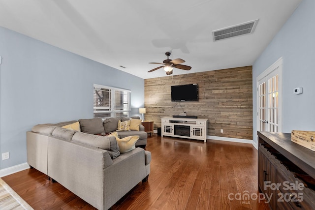 living room featuring an accent wall, wood walls, visible vents, baseboards, and dark wood-style floors