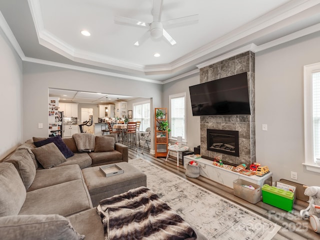 living room with ceiling fan, a tiled fireplace, ornamental molding, wood finished floors, and a raised ceiling