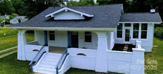 view of front of property featuring covered porch, roof with shingles, a front yard, and brick siding