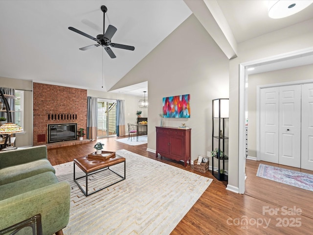 living area featuring wood finished floors, baseboards, high vaulted ceiling, a brick fireplace, and ceiling fan with notable chandelier