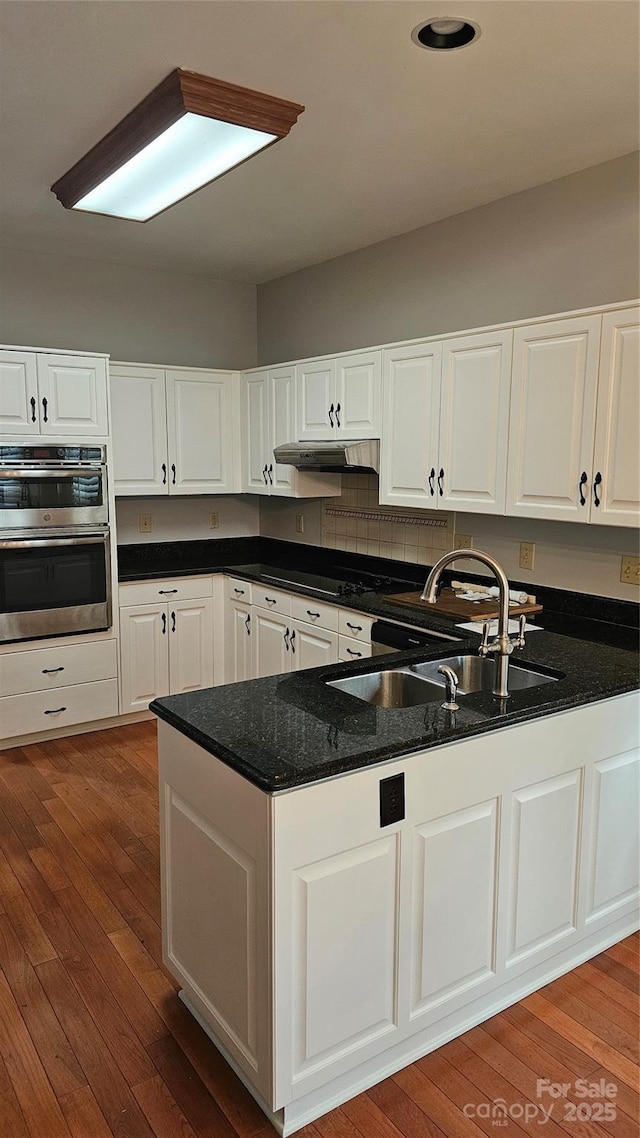 kitchen featuring double oven, white cabinetry, a sink, wood finished floors, and under cabinet range hood