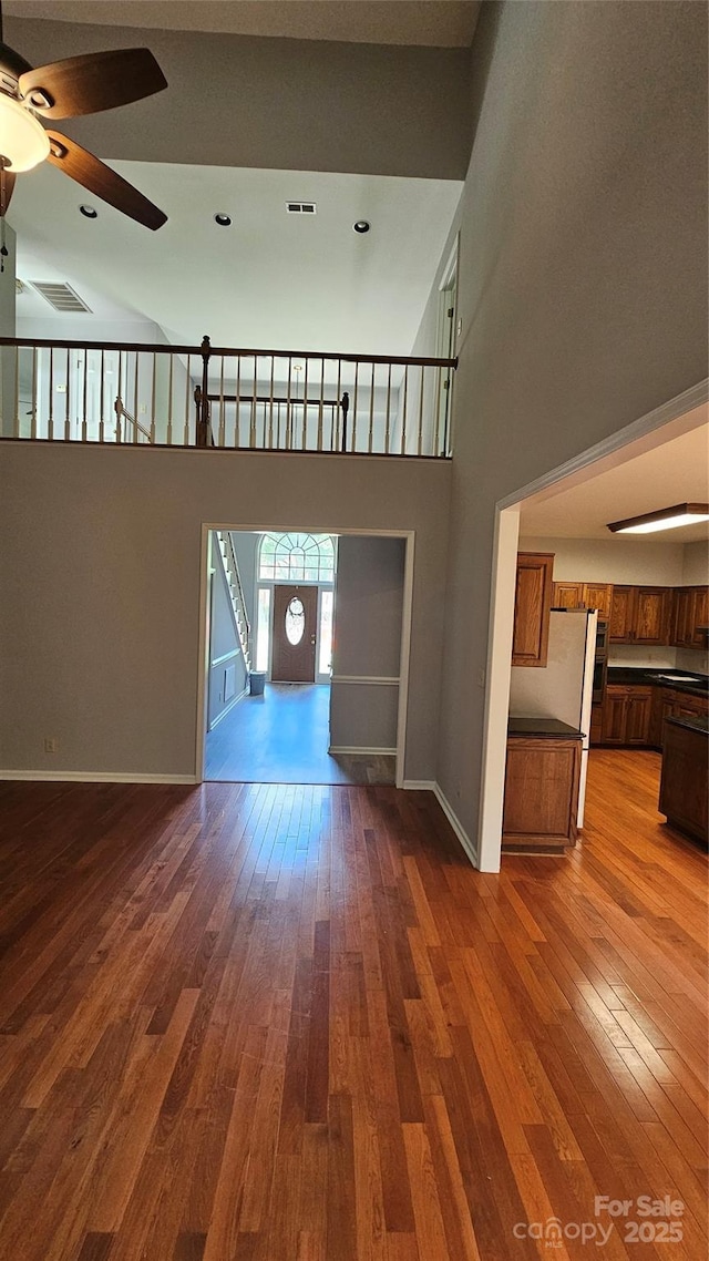 unfurnished living room with wood-type flooring, visible vents, a high ceiling, ceiling fan, and baseboards